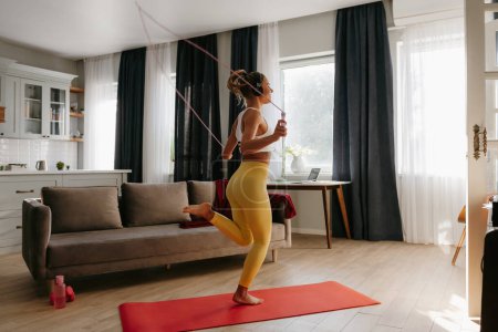 Téléchargez les photos : Pleine longueur de jeune femme sportive dans les écouteurs sautant avec une corde pendant l'entraînement à la maison - en image libre de droit