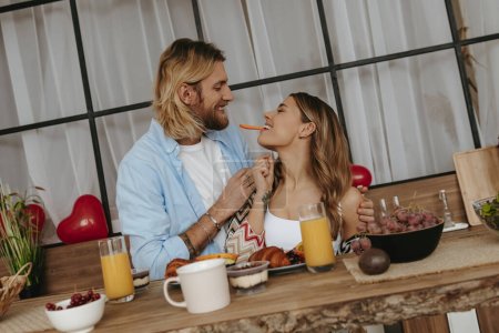 Foto de Juguetona pareja joven disfrutando de un desayuno saludable en casa junto con globos rojos en forma de corazón en el fondo - Imagen libre de derechos