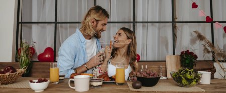 Foto de Feliz pareja romántica disfrutando de un desayuno saludable en casa junto con globos rojos en forma de corazón en el fondo - Imagen libre de derechos