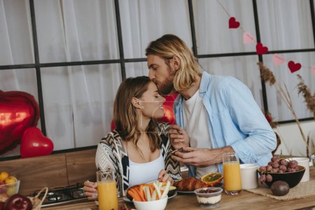 Foto de Romántica pareja joven disfrutando del desayuno en la cocina junto con globos en forma de corazón rojo en el fondo - Imagen libre de derechos