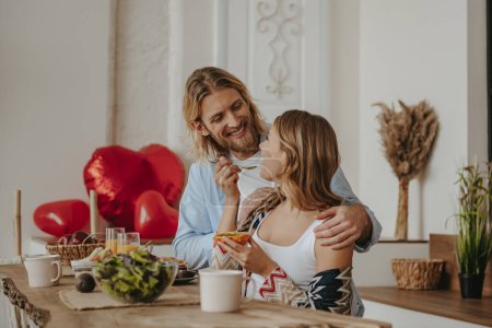 Foto de Joven pareja amorosa disfrutando de un desayuno saludable en casa junto con globos rojos en forma de corazón en el fondo - Imagen libre de derechos