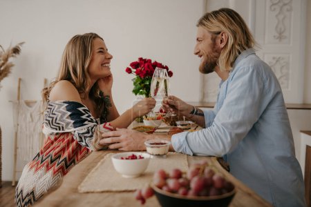 Foto de Alegre joven pareja cariñosa brindis con champán mientras se celebra aniversario en la mesa en casa - Imagen libre de derechos