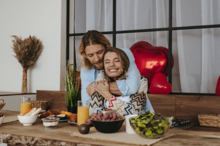 Foto de Joven pareja romántica abrazando mientras se celebra el día de San Valentín en la cocina con globos rojos en el fondo - Imagen libre de derechos