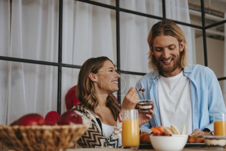 Foto de Mujer joven sonriente alimentando a su novio con el desayuno mientras disfrutan de una comida saludable en la cocina juntos - Imagen libre de derechos