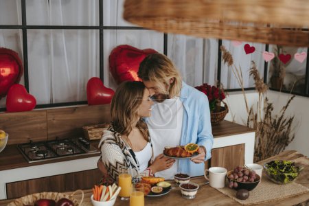 Foto de Sonriente pareja joven desayunando en la cocina junto con globos en forma de corazón rojo en el fondo - Imagen libre de derechos