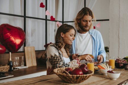 Foto de Hermosa pareja joven desayunando en la cocina junto con globos en forma de corazón rojo en el fondo - Imagen libre de derechos