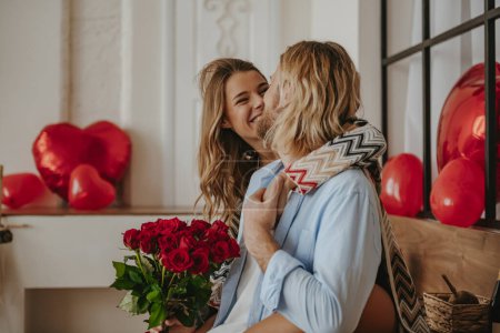 Photo for Young loving couple embracing and holding red roses bouquet while celebrating Valentines day at home - Royalty Free Image