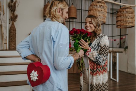 Foto de Joven dando un ramo de flores a su novia sorprendida mientras esconde la caja de regalo detrás de su espalda - Imagen libre de derechos