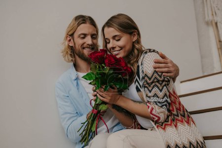 Foto de Romántico joven dando un ramo de rosas a su novia feliz mientras se sienta en las escaleras en casa juntos - Imagen libre de derechos