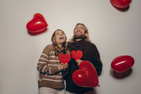 Téléchargez les photos : Joyeux couple amoureux tenant des cartes de Valentines en forme de coeur sur fond beige avec des ballons volant autour - en image libre de droit