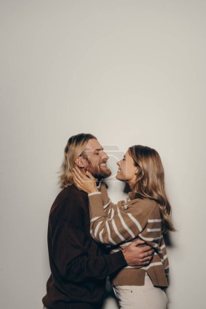 Téléchargez les photos : Joyeux jeune couple aimant dans des vêtements chauds accueillants embrassant face à face tout en se tenant debout sur fond beige - en image libre de droit