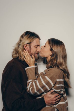 Foto de Feliz joven pareja amorosa en abrigo cálido acogedor abrazar y besar, mientras que de pie sobre fondo beige - Imagen libre de derechos