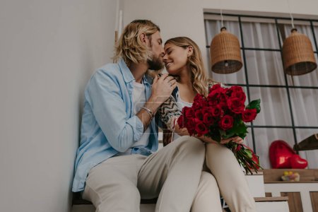 Foto de Joven pareja romántica abrazando y besándose en casa mientras mujer sosteniendo ramo de rosas rojas - Imagen libre de derechos