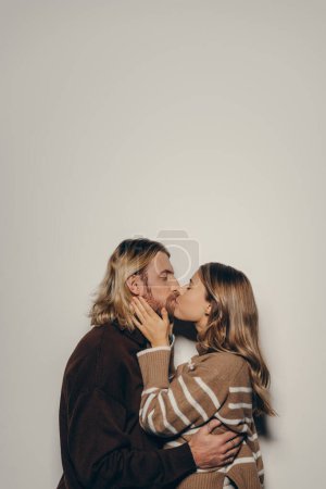 Téléchargez les photos : Romantique jeune couple amoureux dans des vêtements chauds accueillants embrassant et embrassant tout en se tenant debout sur fond beige - en image libre de droit