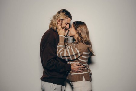 Téléchargez les photos : Joyeux jeune couple aimant dans des vêtements chauds accueillants embrassant face à face tout en se tenant debout sur fond beige - en image libre de droit