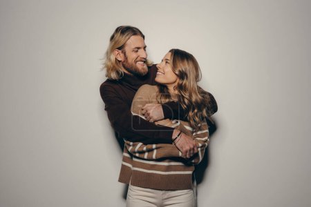 Foto de Alegre joven amante pareja en acogedor cálido desgaste abrazando mientras de pie sobre fondo beige - Imagen libre de derechos