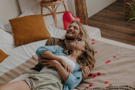 Foto de Feliz pareja cariñosa abrazando y divirtiéndose mientras está acostada en la cama rodeada de globos rojos en forma de corazón - Imagen libre de derechos