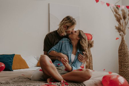 Foto de Romántica pareja amorosa sentada en la cama y besándose mientras el corazón rojo forma confeti volando alrededor - Imagen libre de derechos