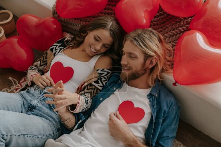 Foto de Vista superior de pareja amorosa sosteniendo tarjetas de San Valentín en forma de corazón mientras se apoya en la cama rodeada de globos rojos - Imagen libre de derechos