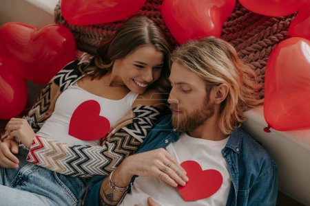 Téléchargez les photos : Vue du dessus du couple romantique tenant des cartes de Saint-Valentin en forme de coeur tout en étant couché sur le lit entouré de ballons rouges - en image libre de droit