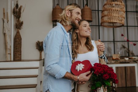 Téléchargez les photos : Jeune couple romantique embrassant et tenant boîte cadeau et fleurs tout en célébrant l'anniversaire à la maison - en image libre de droit