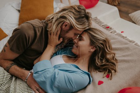 Foto de Joven pareja cariñosa abrazándose y besándose mientras está acostada en la cama rodeada de globos rojos en forma de corazón - Imagen libre de derechos