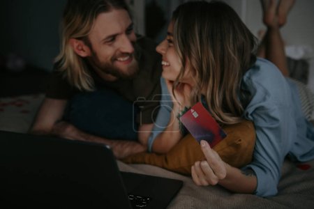 Photo for Joyful loving couple lying in bed while using laptop and credit card for online shopping from home - Royalty Free Image