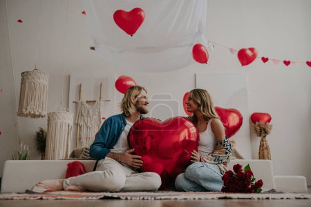 Foto de Feliz pareja amorosa sosteniendo gran globo en forma de corazón rojo mientras se celebra el día de San Valentín en casa acogedora - Imagen libre de derechos