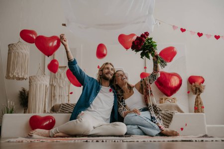 Photo for Happy loving couple celebrating Valentines day at home together with red heart balloons flying on background - Royalty Free Image