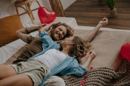 Foto de Relajada pareja cariñosa abrazando y sonriendo mientras está acostado en la cama rodeado de globos rojos en forma de corazón - Imagen libre de derechos
