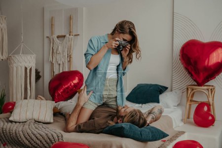 Téléchargez les photos : Jeune femme photographiant son petit ami avec une caméra rétro tout en se relaxant au lit à la maison ensemble - en image libre de droit