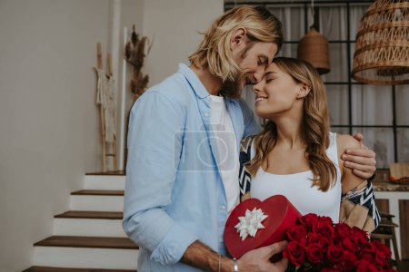 Téléchargez les photos : Jeune couple romantique embrassant et tenant boîte cadeau et fleurs tout en célébrant l'anniversaire à la maison - en image libre de droit