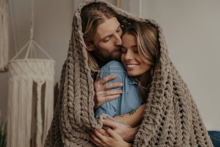 Photo for Young loving couple covering with a cozy warm blanket while spending romantic time at home together - Royalty Free Image