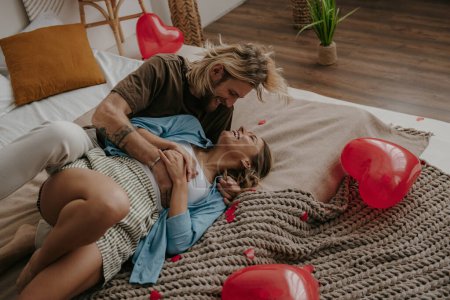 Téléchargez les photos : Couple aimant ludique embrassant et s'amusant tout en étant assis sur le lit entouré de ballons rouges en forme de coeur - en image libre de droit