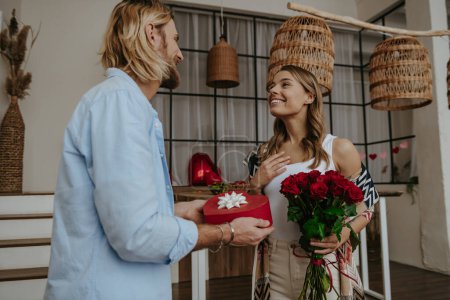Photo for Handsome young man giving a flower bouquet and gift box to his surprised girlfriend standing at home - Royalty Free Image