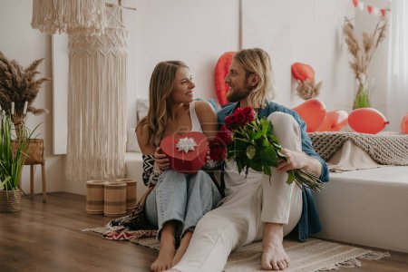 Photo for Loving couple embracing and holding roses and gift box while spending Valentines Day at cozy home - Royalty Free Image