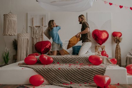 Téléchargez les photos : Heureux jeune couple aimant avoir une bataille d'oreillers dans le lit entouré de ballons rouges en forme de coeur - en image libre de droit