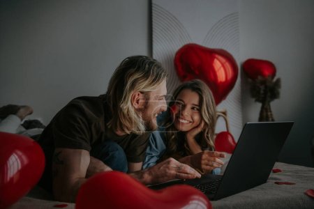 Foto de Alegre pareja amorosa utilizando el ordenador portátil y compras en línea en el día de San Valentín desde el dormitorio decorado - Imagen libre de derechos
