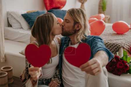 Téléchargez les photos : Couple aimant souriant embrassant et embrassant tout en tenant des cartes Valentines en forme de coeur dans la chambre décorée - en image libre de droit