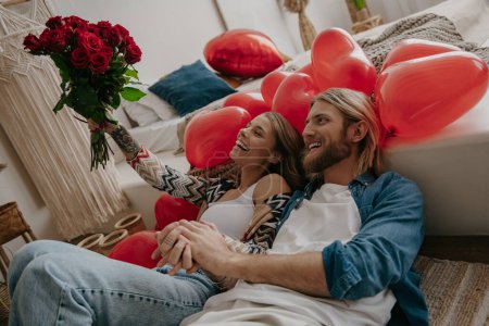Foto de Feliz pareja amorosa apoyada en la cama rodeada de globos rojos mientras la mujer sostiene un ramo de rosas rojas - Imagen libre de derechos