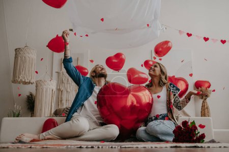 Foto de Pareja sonriente sosteniendo globo en forma de corazón rojo y lanzando confeti mientras celebra el día de San Valentín en casa - Imagen libre de derechos