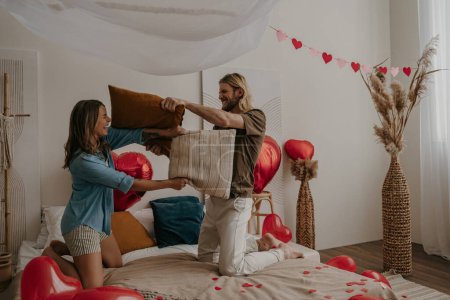 Foto de Juguetona pareja amorosa teniendo una pelea de almohadas en la cama rodeada de globos rojos en forma de corazón - Imagen libre de derechos