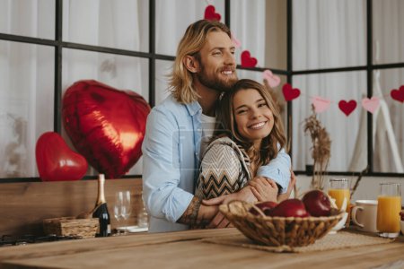 Foto de Sonriente pareja joven abrazando mientras se celebra el día de San Valentín en la cocina con globos rojos en el fondo - Imagen libre de derechos