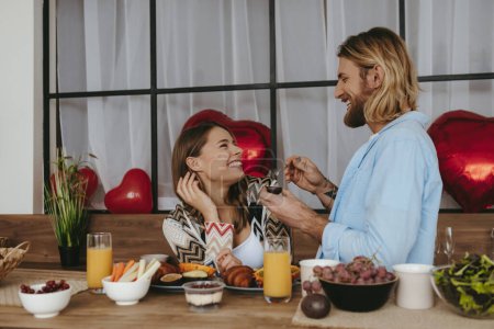 Téléchargez les photos : Souriant jeune homme nourrir sa petite amie avec le petit déjeuner tout en appréciant la nourriture saine à la cuisine ensemble - en image libre de droit