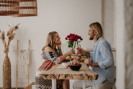 Foto de Joven pareja alegre y cariñosa brindando con champán mientras cenan románticamente en casa de estilo boho - Imagen libre de derechos