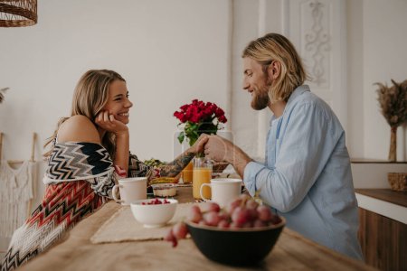 Téléchargez les photos : Romantique jeune couple tenant la main tout en appréciant le petit déjeuner sain à la maison de style boho - en image libre de droit