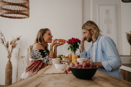 Foto de Joven pareja alegre disfrutando de comida y bebidas saludables mientras desayuna en casa de estilo boho - Imagen libre de derechos
