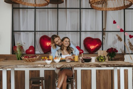 Foto de Sonriente pareja joven abrazando mientras se celebra el día de San Valentín en la cocina con globos rojos en el fondo - Imagen libre de derechos