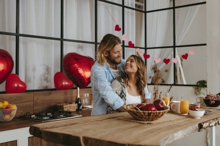 Foto de Feliz pareja joven abrazando mientras se celebra el día de San Valentín en la cocina con globos rojos en el fondo - Imagen libre de derechos