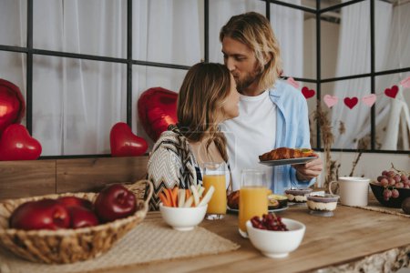 Foto de Sonriendo joven pareja besándose mientras desayuna en la cocina junto con globos rojos en el fondo - Imagen libre de derechos